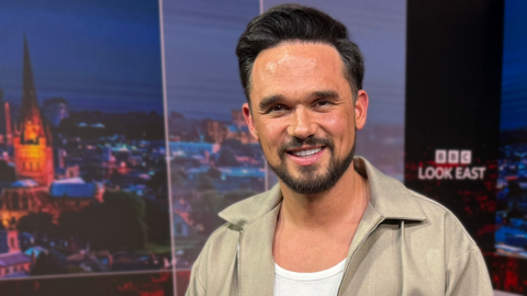 A head and shoulders image of a man smiling at the camera in a TV news studio. He has short dark hair, a trimmed beard and is wearing a white top with a beige unzipped collared shirt over the top. 