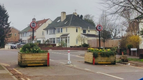 Image shows a road with two large wooden square planters filled with flowers either side of a white bollard.