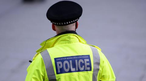 A Greater Manchester Police officer stands with his back to the camera, wearing a hi-vis jacket and police cap, a logo on the back, in blue with white font says 'Greater Manchester Police'