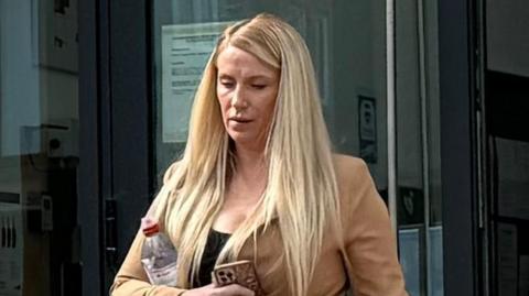 A woman with long blonde hair tied up in a ponytail walks out of court looking down. She wears a black top with a beige jacket. She has a water bottle tucked under her arm and is holding an iPhone.
