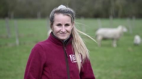 Heidi Holder is in a field. Grass and sheep are in the background. Heidi is wearing a maroon jacket and has her hair tied up in a ponytail.