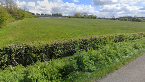 A large grass field viewed from the edge of a country road, with a trimmed hedgerow running horizontally across the front of the image, separating the roadside verge from the field.