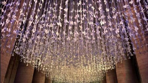 Thousands of paper doves hang from the ceiling of an abbey with purple lighting shining over them.