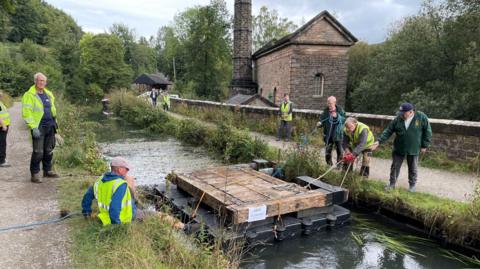 A wooden lock gate on a floating pontoon on a canal, which has ropes attached and is being towed by six volunteers. 