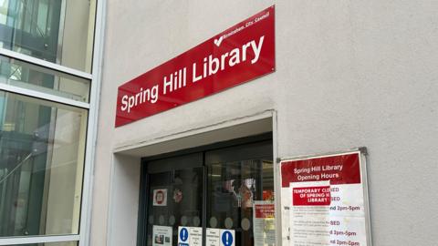 The sign for Spring Hill Library, a Birmingham City Council building. It has a modern door and its opening hours are taped over by a closure notice. 