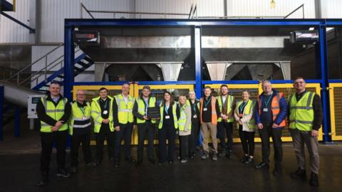 Ashley Nicholson, chief business officer at Port of Tyne and Sean Coyle, CEO, Origin Enterprises alongside employees from Port of Tyne and Origin Soil Nutrition. They stand in front of a yellow metal fence separating them from a large metal machine.