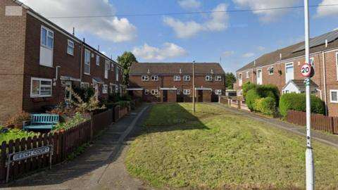 A small court area of brown brick terraced houses overlooking a grass area in the centre