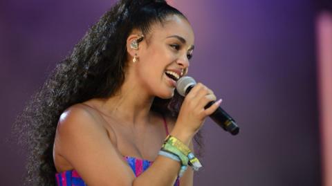 A woman with black curly hair and wearing a pink and blue top singing into a microphone 