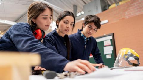 Stock photo shows three students looking down at a workbench with a protective mask on the table, wearing overalls and earmuffs in a design technology schooling environment.