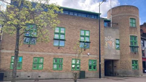 A brown-bricked building with green-framed windows. The words "CROWN COURT" are written above the door. Two trees can been seen in a paved area at the front of the building.
