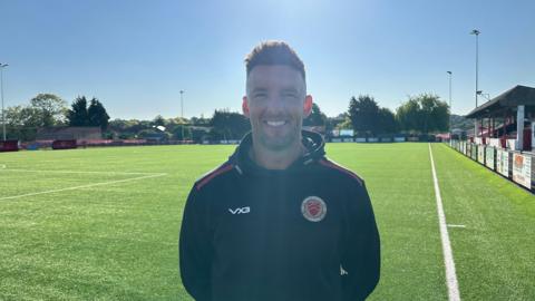 Jamie Coyle looks at the camera and smiles. He is standing on a football pitch and wearing a Whitstable training jacket.