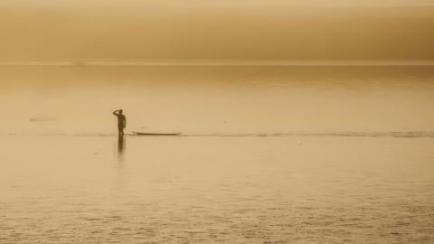 A surfer salutes on the still water at dusk as he stands next to his surfboard. The sky and the river appear orange due to the haze.