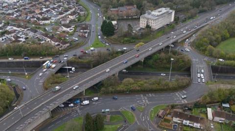An ariel view of Manadon roundabout in Plymouth. There is a large area at the centre with trees either side of it. There are cars driving on the road and housing in the top left corner. There are two larger buildings to the right of the houses.