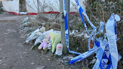 Flowers and a teddy bear rest near a lamppost, with bunched-up blue-and-white police tape lying to the side