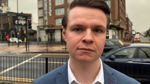 A serious-looking man wearing a white shirt and a blue blazer stood next to a busy road 