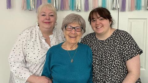 Anne is stood in the middle of her daughter Donna and her granddaughter Sarah. Anne has short grey hair and is wearing dark rimmed glasses, a blue jumper and a gold necklace and earrings. Donna has short blonde hair and is  wearing a white blouse with black hearts on and gold hoop earrings. Sarah has dark hair and is wearing a floral dress.