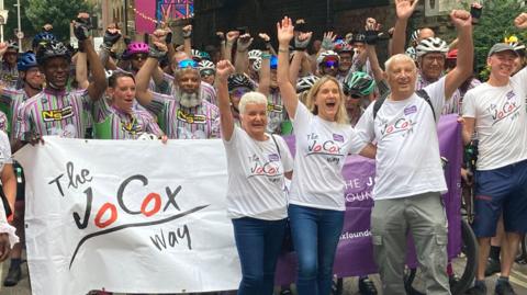 A group of people with their arms held aloft. Some of them are wearing white T-shirts and others are holding a white flag that reads "The Jo Cox Way".