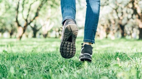 Person walking on grass. Only lower legs and feet in shot, with little blue jeans and grey, white and black trainers