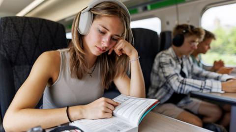 GCSE Biology student on a train with headphones and a text book, listening to a BBC Bitesize revision podcast.
