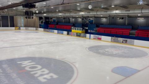 The inside of the ice rink - it has ice, surrounded by white boarding making the edges. There are red seats behind the boarding.