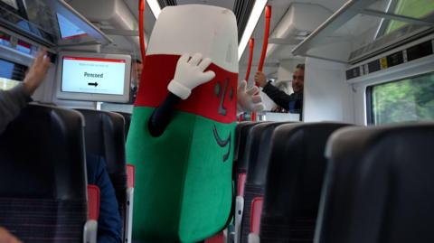 The red, green and white triangle character aboard the new Transport for Wales train. He is standing in the aisle with his hands in the air. 