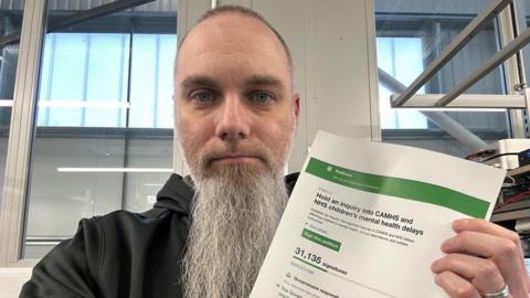 Tom is holding a petition in his hands and wearing a black hoodie. 