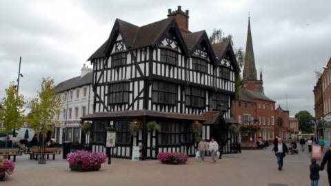 A black-and-white Jacobean timber-framed house, Old House Museum, in Hereford. It is surrounded by planters full of colourful flowers and there is a church spire in the background.