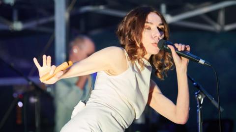 PJ Harvey singing into a microphone. She has shoulder length curly brown hair and is wearing a fitted white dress.