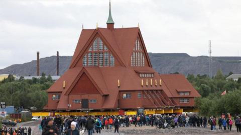 A church - a vast red timber structure - is hoisted on giant trailers and is being rolled along a road.  Large crowds are gathered around the church. 