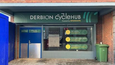 The outside of Derbion Cycle Hub. It has a blue double door on the left, window on the right and a sign above on a green background which reads "Cyclehub"