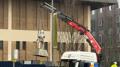A vehicle lifts up a metal statue of a steel worker with its crane. A worker can be seen operating the crane on the right.