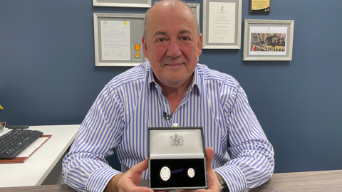 Phil McDermott, in a blue striped shirt holding an Elizabeth Emblem medal in a case. He is sitting in an office with blue walls and a computer to his right