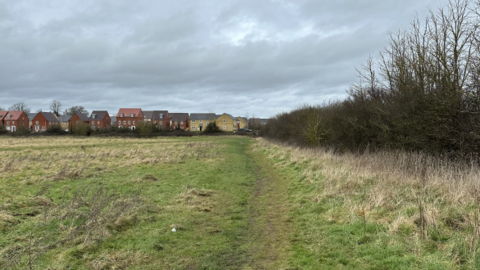 Image shows a field with a trodden pathway down the side. There are red brick houses at the back of the field.