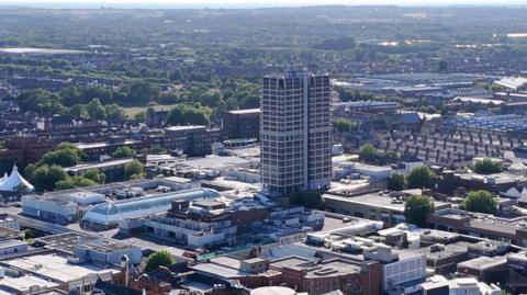 An overhead view of a densely built town centre with a tall tower block in the centre of the frame, as well as the glass roof of a large shopping centre, while greenery stretches into the distance.