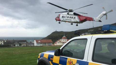 A red and white helicopter takes off from a field in Port Erin. You can see houses, hills and the sea in the distance. A 4x4 vehicle can be seen in the foreground. It is white and has a blue and yellow checkerboard pattern and the government crest along the side