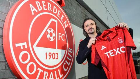 Lutz Pfannenstiel holds up an Aberdeen shirt while standing next to a large sign bearing the club's logo