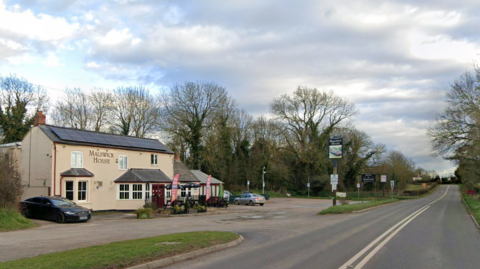 A two-storey pub with cream rendering sits alongside a B-road. There is a large car park at the front of the pub. The road is empty, with trees lining either side of it. It is a grey day.