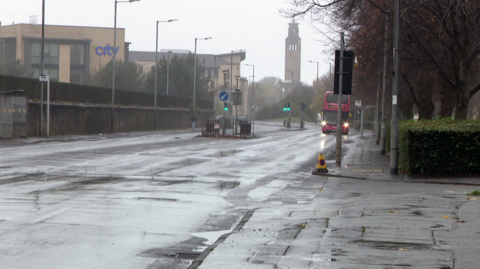 Junction of road on a grey, rainy day. There is a red bus approaching with headlights on and a tower in the distance 