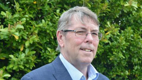 Nick Thulbourn stares off the camera to the right. He wears glasses and a blue suit with the top button of his shirt undone. The background is a green bush.