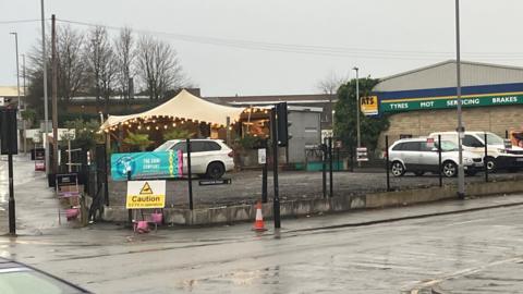 A car park in Bradford, with a few cars parked in unmarked bays. The car park is next to an ATS garage, with a canopy and a small cafe-type hut in the distance. Roads surround the car park on all sides. 