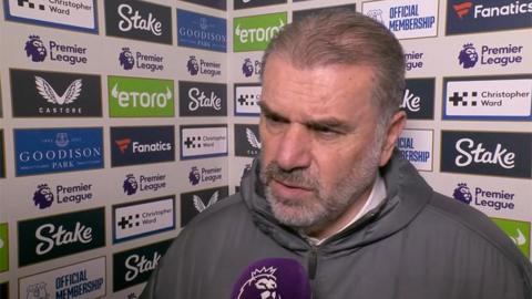 Ange Postecoglou, wearing a grey jacket, speaking to a reporter holding a purple microphone. Behind him is a sponsors board.