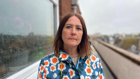 Katie Courtney-Jones standing outside flats in Guildbourne Court on a walkway on the top floor next to a window. She has brown hair and a flowery rain coat