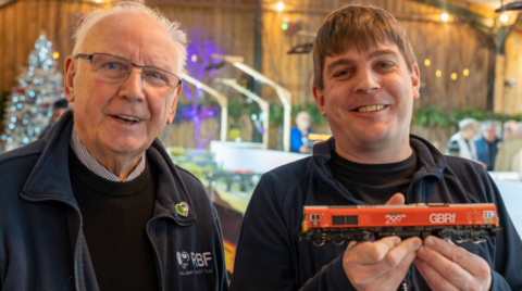 Two men in dark clothing are next to each other looking at the camera. The man on the right is holding a model train. 