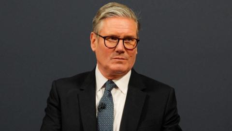 Sir Keir Starmer has grey hair which is slicked back to one side of his head with shorter sides and is wearing oval glasses, a black suit jacket, white shirt and blue tie with yellow triangular print on it. He has a microphone attached to his tie and is looking to the side with a serious expression. The background is dark grey.