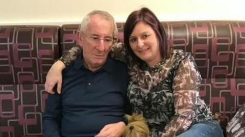 Michael Griffiths, with short white hair and glasses, wearing a blue sweater, is sitting alongside Brenda Brereton, who has long brown hair and is wearing a brown and white patterned top. She has her right arm around her father's shoulders. They are sitting on a brown sofa with a red and white pattern.