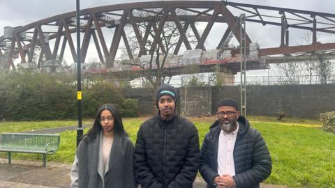 Three people wearing dark clothing stand together in a line looking at the camera. There is a woman and two men. Behind them is a huge steel structure which is one of the HS2 viaducts.