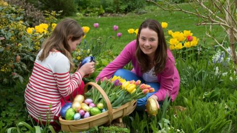 Two girls are sat on some grass filling a basket with flowers and foil-wrapped chocolate eggs of various colours. 