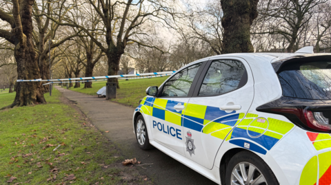 Police car on path in park with cordon beyond and trees lining path