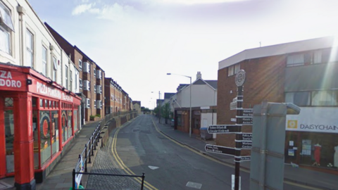 Street view of Mill Street in Evesham. There is a red pizza restaurant at the end of the street and a row of flats down the left side of the street.