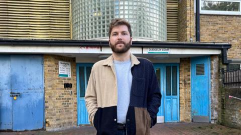 An image of Max Templer, a young man with short brown hair and a beard wearing a patchwork jacket and grey T-shirt. He is stood in front of the blue front doors to his block of flats